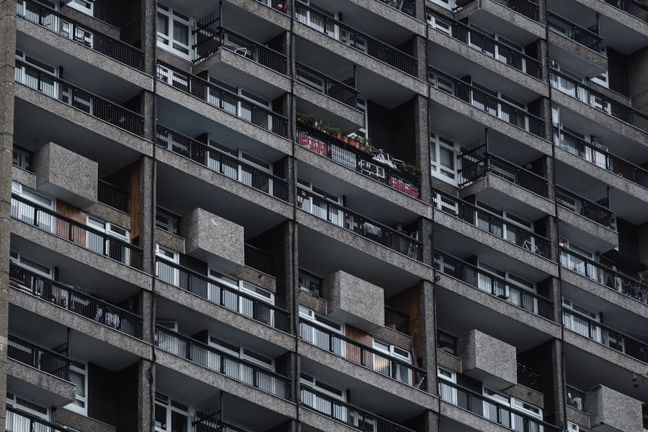 A close-up view of a multi-story residential building with numerous balconies featuring black metal railings and glass paneling, each with personal outdoor decorations such as potted plants and laundr