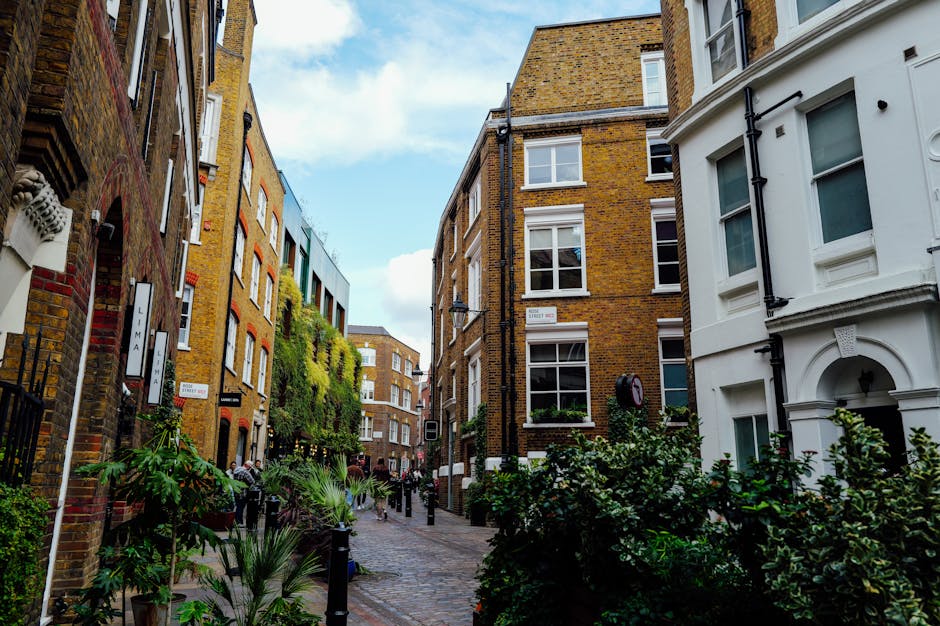A narrow cobblestone street lined with multi-story brick and white-painted buildings, some with large windows and decorative architectural details. The buildings are closely spaced, creating a cozy ur